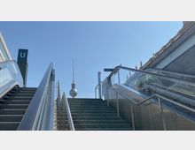 Blick von unten auf eine Treppe und eine Rolltreppe an einem U-Bahneingang, mit dem Fernsehturm im Hintergrund und einem Gebäude mit Statuen am rechten Bildrand unter blauem Himmel.
