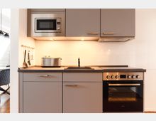 Moderne Einbauküche mit grauen Schränken, einer Arbeitsfläche aus Holzoptik, integriertem Herd und Backofen sowie einer Mikrowelle. Die Wand ist mit weißen Fliesen verkleidet, und Kochutensilien hängen an einer Stange neben einem Wasserhahn mit Spülbecken.