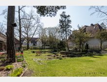 Ein gepflegter Garten mit einer Rasenfläche, Blumenbeeten und vereinzelten Bäumen, umgeben von Häusern mit Satteldächern. Im Vordergrund stehen blühende Pflanzen und ein Baumstumpf, während sich im Hintergrund weitere Bäume und Hecken befinden.