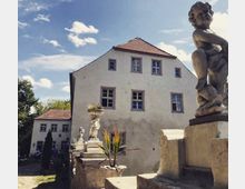 Ein historisches Herrenhaus mit hellen Putzfassaden und roten Ziegeldächern, umgeben von dekorativen Steinstatuen und Pflanzgefäßen. Im Vordergrund ist eine Balustrade mit barocken Verzierungen erkennbar, während im Hintergrund Bäume und ein Hofbereich zu sehen sind.