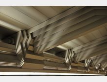 Decke mit geometrischen, schichtartig angeordneten Strukturen aus Metallpaneelen, beleuchtet durch eine Lichtquelle, die weiche Schatten wirft.