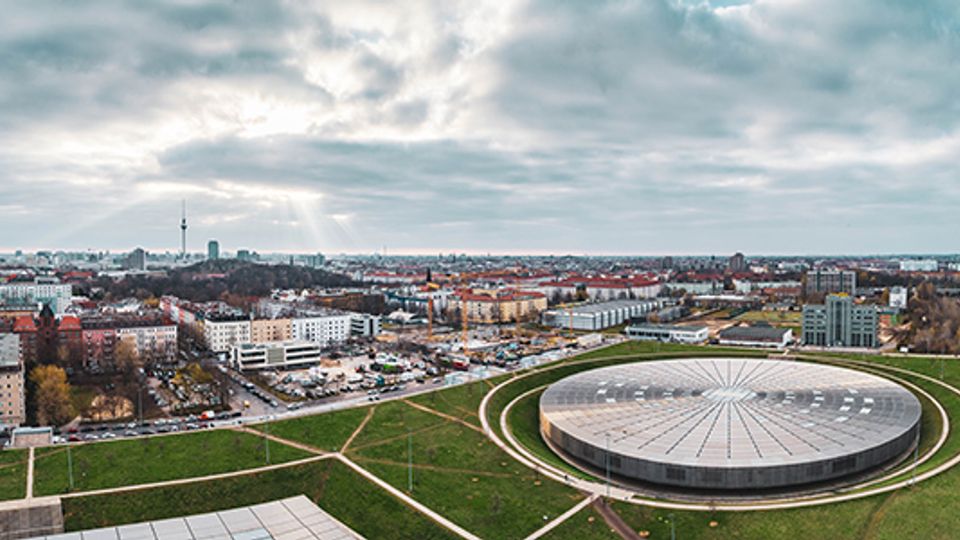 Panoramablick über eine weitläufige Stadtlandschaft mit dicht bebauten Wohn- und Gewerbegebieten unter bewölktem Himmel. Im Vordergrund liegt eine große, runde Arena mit flachem, radial gegliedertem Dach, umgeben von Grünflächen und Wegen.