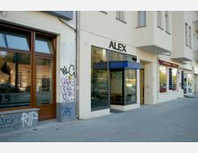 Straßenansicht eines Gebäudes mit Geschäften im Erdgeschoss. Zu sehen sind ein Eingang zu einem Hotel mit dem Namen „Alex“, große Schaufenster und Fassaden mit Graffiti, daneben weitere Geschäftseingänge.