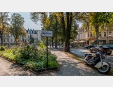 Straßenszene am Karlsplatz mit Kopfsteinpflasterwegen, umgeben von historischen Gebäuden mit verzierten Fassaden und einem kleinen grünen Parkbereich. Im Vordergrund steht ein Schild mit der Aufschrift "Karlsplatz", dahinter eine geparkte Motorräder und Autos sowie ein Café mit Sonnenschirmen.