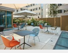 Offener Innenhof mit Sitzgelegenheiten, bestehend aus modernen Tischen und bunten Stühlen. Die Umgebung ist von Pflanzen, einer Holzwand und umliegenden Bürogebäuden eingerahmt, mit großen Sonnenschirmen, die Schatten spenden.