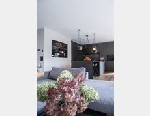 Moderne Wohnküche mit dunklem, minimalistischem Küchenbereich und einer zentralen Kücheninsel, darüber hängende Lampen mit schlichten Kabeln. Im Vordergrund ein graues Sofa und ein Blumenstrauß aus getrockneten Hortensien; Holzfußboden und dezente Wanddekoration.