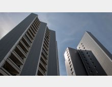 Hochhäuser mit grauen Fassaden und Balkonen, aufgenommen aus einer Froschperspektive gegen einen leicht bewölkten Himmel. Beide Gebäude haben eine moderne, geometrische Architektur mit mehreren Stockwerken und symmetrischen Fenstern.