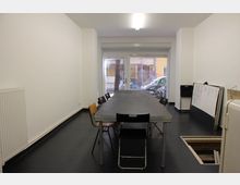 Ein schlichter Besprechungsraum mit einem langen rechteckigen Tisch, umgeben von schwarzen und holzfarbenen Stühlen. Im Hintergrund befindet sich ein großes Fenster mit Blick auf eine Straße, und an der rechten Wand sind eine Tafel und diverse Gegenstände angelehnt.