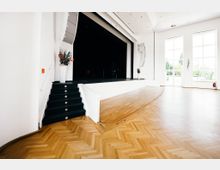Heller Veranstaltungssaal mit einer erhöhten Bühne, schwarzer Treppe und hellem Holzparkettboden. Große Fenster auf der rechten Seite lassen Tageslicht herein, dekoriert mit weißen Wänden und Blumenschmuck.