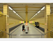 U-Bahn-Station mit gelben Kacheln und linearen Beleuchtungselementen; ein stehender U-Bahn-Zug der Linie U5 befindet sich rechts, während Personen vor einem Infopunkt stehen. Die Station ist mit der Beschriftung "Weberwiese" gekennzeichnet, und zentrale Pfeil- und Hinweiszeichen hängen von der Decke.