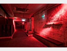 Ein Kellerraum mit grauen Betonwänden, schwach beleuchtet durch rote Lichter. An den Wänden befinden sich Graffiti, ein beleuchtetes Notausgangsschild und eine gemalte Szene auf einem Vorhangmotiv, während Holzpaletten an den Seiten gestapelt sind.