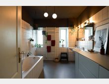 Helles Badezimmer mit weißen Fliesen, einer Badewanne, zwei Fenstern, einem Doppelwaschbecken und einem großen Spiegel. Pflanzen und warme Beleuchtungselemente schaffen eine gemütliche Atmosphäre, während Handtücher an einem Heizkörper hängen.