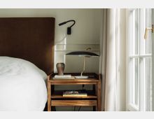 Ein minimalistisches Schlafzimmer mit einem braunen Kopfteil, einem Nachttisch aus Holz, darauf eine Tischlampe mit schwarzem Schirm und goldfarbenem Fuß, sowie ein paar Bücher und eine Keramikvase. Rechts befindet sich ein bodentiefes Fenster mit weißen Vorhängen.