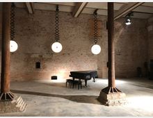 Industriehalle mit unverputzten Backsteinwänden, freiliegenden Holzdeckenbalken und vier Metallpfeilern. Im Raum steht ein Flügel mit schwarzer Abdeckung, beleuchtet durch drei runde Deckenlampen, die an gefederten Halterungen hängen.