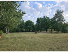 Weitläufige Wiese mit vereinzelten Bäumen und einem kleinen Spielgerüst aus Holz. Im Hintergrund sind dichte Bäume und ein alter landwirtschaftlicher Pflug erkennbar, darüber ein teilweise bewölkter Himmel.