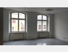 Leerer Raum mit hellgrauem Boden und zwei großen, bogenförmigen Fenstern, die Tageslicht hereinlassen. Unter den Fenstern befinden sich Heizkörper; die Wände sind weiß gestrichen, und die Decke ist mit rechteckigen Paneelen abgehängt.