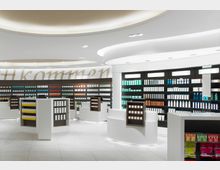 Ein moderner Apotheken-Innenraum mit weißen Regalen und geschwungener Beleuchtung an der Decke. Die Regale sind ordentlich mit Medikamenten und Pflegeprodukten in verschiedenen Farben bestückt; zentral stehen freistehende Ausstellungsregale.