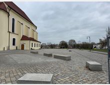 Platz mit Kopfsteinpflaster und modernen Betonelementen als Sitzgelegenheiten, neben einer großen, gelb gestrichenen Kirche mit rotem Dach. Im Hintergrund sind Bäume, Straßenlaternen und vereinzelte Gebäude zu sehen.