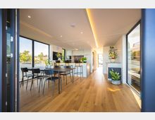 Modernes Wohn- und Esszimmer mit großen bodentiefen Fenstern, die einen Blick auf eine Wasserlandschaft bieten. Der Raum ist mit hellen Holzböden, einem Esstisch mit schwarzen Stühlen, einer Küchenzeile mit Barhockern und mehreren dekorativen Pflanzen gestaltet.