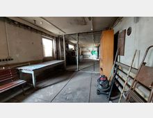 Ein industrieller oder leerstehender Raum mit einfachen Möbeln, darunter ein Holz- und Metalltisch sowie eine Sitzbank. Im Hintergrund sind große Fenster, metallene Strukturen, ein offenes Regal und ein grünes Schalttafel-Panel zu sehen.