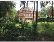 Einfamilienhaus mit orangefarbener Fassade und Satteldach, umgeben von einem großen Garten mit blühenden Rosen, Rhododendren und hohen Bäumen. Der Garten wirkt gepflegt, mit einem Rasenbereich und vereinzelten Sitzgelegenheiten.