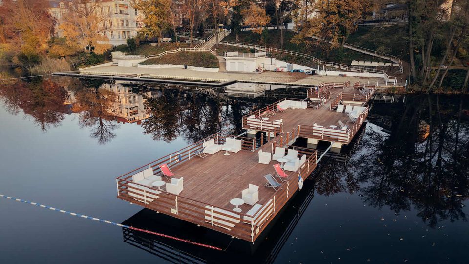 Floating wooden dock platforms on a calm lake, furnished with white seating and red lounge chairs and bordered by railings. In the background, a terraced shoreline with stairways leads up to trees with autumn foliage and a large multi-story building reflected in the water.