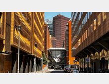 Straßenansicht am Potsdamer Platz in Berlin mit modernen Gebäuden aus orangefarbenen Klinkern und Glasfassaden. Im Hintergrund ragt ein rotes Hochhaus auf, und in der Mitte ist ein Eingang mit der Beschriftung "Potsdamer Platz" zu sehen.