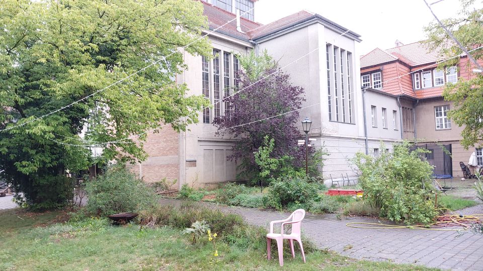 Außenbereich eines Gebäudekomplexes mit grauer und beiger Fassade, großen Fenstern und einem roten Ziegeldach. Der Garten ist von Bäumen, Büschen und Rasenflächen geprägt, mit einem gepflasterten Weg, einem rosa Plastikstuhl und einem Holzgestell im Hintergrund.