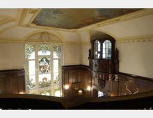 Innenansicht eines historischen Gebäudes mit dekorativen Buntglasfenstern, dunkler Holzvertäfelung und einer kleinen Holzempore an der Wand. Die Decke ist mit kunstvollen Wandmalereien verziert.