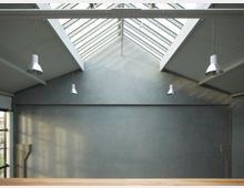 Innenraum eines modernen Gebäudes mit hohen grauen Wänden, einer schrägen Decke aus Glaspaneelen und freiliegenden Metallträgern. Vier Hängelampen aus Metall sind von der Decke herabgehängt, der Raum wirkt offen und minimalistisch.