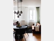 Wohnzimmer mit moderner Einrichtung: ein runder, schwarzer Esstisch mit mehreren schwarzen Stühlen, ein braunes Ledersofa mit weißer Decke, ein Sessel aus Leder und ein schwarzer Teppich. Im Hintergrund helle Vorhänge, zwei große Fenster und eine Hängelampe mit mehreren Kugel-Leuchten.