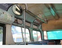 Innenraum eines alten Linienbusses mit gepolsterten Sitzen, großen Fenstern und einer abgerundeten Decke. Sichtbare Details umfassen eine Sicherheitsanweisung auf Englisch und Spanisch sowie Metallstangen und einen Rückspiegel.