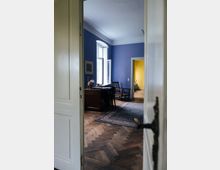 Blick durch eine offene Tür in einen Altbau-Raum mit blau gestrichenen Wänden, Stuckdecke und Fischgrät-Parkett. Am hohen Fenster stehen ein Schreibtisch und ein Stuhl, ein gemusterter Teppich liegt auf dem Boden, und durch eine weitere Tür ist ein gelb gestrichener Nebenraum zu sehen.