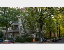 Ecke einer Wohnstraße mit einem mehrstöckigen Gebäude im Altbaustil, umgeben von dichtem Baumbewuchs. Auf der Straße stehen Autos und ein Müllcontainer, während der Gehweg von Hecken gesäumt ist.