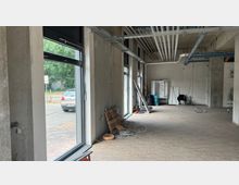 Ein leerer Innenraum im Rohbau mit großen Fenstern, durch die eine Straße und Bäume sichtbar sind. Die Decke zeigt freiliegende Lüftungsrohre und Kabel, während Baumaterialien und Werkzeuge auf dem Boden verteilt sind.