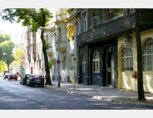 Straßenszene in einem Wohngebiet mit mehrstöckigen Gebäuden, die eine helle, teils gelbliche und dunkle Fassade mit Bögen und Fenstern haben, Bäume entlang des Gehweges und parkende Autos am Straßenrand. Ein Haltestellenschild und ein Verkehrsschild sind sichtbar.