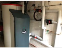 Heizungsraum mit einem großen weißen Warmwasserspeicher und einem grauen Heizungsmodul. An der Wand verlaufen mehrere isolierte Rohre, ein Heizungsregler und Absperrventile.