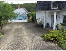 Gepflasterter Hofbereich vor einem weißen Haus mit schwarzem Schrägdach und Balkon, umgeben von blühenden Pflanzen in Töpfen. Im Hintergrund eine Wand mit einem Wandgemälde, das eine ländliche Szene mit Wasser, einer Hütte und einem Steg darstellt, sowie Bäume und Büsche im Umfeld.