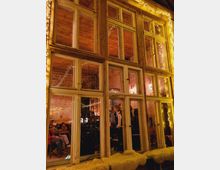 Holzgerahmte Fenster eines Gebäudes mit Blick in einen gemütlich beleuchteten Innenraum, in dem Personen an Tischen sitzen und eine Person an einem Mikrofon steht; Lichterketten schmücken den Raum und das Fenster.