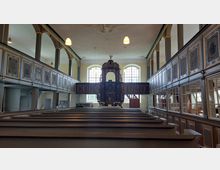 Innenraum einer historischen Kirche mit Holzbänken im Vordergrund, einer reich verzierten Kanzel in der Mitte und umlaufenden Emporen mit bemalten Feldern. Große Fenster lassen Tageslicht herein, und die Decke ist mit einfachen Stuckelementen dekoriert.