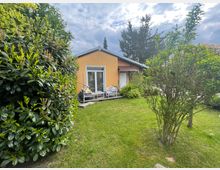 Ein kleines, einstöckiges Haus mit gelber Fassade und einem roten Ziegeldach, umgeben von einem gepflegten Garten mit Rasen, Sträuchern und Bäumen. Vor dem Haus befindet sich eine Terrasse mit weißen Gartenmöbeln.