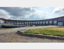 Historischer Rundlokschuppen mit mehreren Toren aus rotem Backstein und großflächigen Fenstern, im Vordergrund eine Drehscheibe für Züge sowie gepflasterte Wege und eine Grünfläche. Der Himmel ist leicht bewölkt.