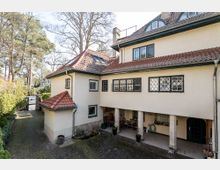 Zweistöckiges Wohnhaus mit hellbeiger Fassade und roten Ziegeldächern, großen Fenstern mit Gitterverzierungen und überdachtem Eingangsbereich mit Säulen. Umgeben von einem gepflasterten Hof, Bäumen und Hecken.