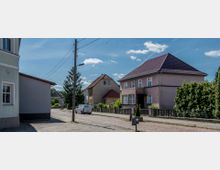 Eine ruhige Wohnstraße mit gepflastertem Gehweg, umgeben von Einfamilienhäusern mit Satteldächern. Im Vordergrund ist ein helles Haus mit Vorgarten und einem parkenden Transporter zu sehen, während Strommasten die Straße säumen.