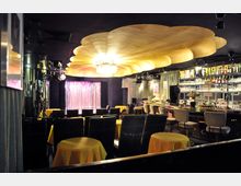 Innenraum eines eleganten Nachtclubs mit dunklem Mobiliar. Auffällig ist eine große, goldfarbene Deckeninstallation in Form einer Muschel, darunter eine Bühne mit glitzerndem Vorhang, sowie eine gut bestückte Bar mit gepolsterten Barhockern.