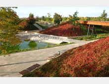 Ein moderner Park mit terrassierten Grünflächen, roten Bodendeckern und Bäumen, einer kleinen Wasserfläche mit Stegen und Geländern sowie einer erhöhten, rostfarbenen Fußgängerbrücke. Im Hintergrund sind Treppen und weitere Pflanzen sichtbar.