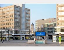 Ansicht des Alexanderplatzes in Berlin mit modernen Gebäuden, großen Glasfenstern und einem U-Bahn-Eingang mit einem blauen Schild "U Alexanderplatz" im Vordergrund. Im Hintergrund ist die Kuppel des Berliner Doms zu sehen.