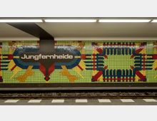 U-Bahnhof mit farbenfrohen Mosaiken an der Wand, der Schriftzug "Jungfernheide" ist auf einem braunen Schild abgebildet. Die Wand zeigt geometrische, abstrakte Muster aus grünen, gelben, roten und blauen Fliesen, beleuchtet durch Deckenleuchten.