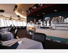 Innenansicht eines modernen Restaurants mit Panoramafenstern, die viel Tageslicht hereinlassen. Im Vordergrund sind gedeckte Tische und schwarze Stühle zu sehen, im Hintergrund eine Theke mit Kaffeemaschinen und Tassen, dekoriert mit hängendem Weihnachtsschmuck.
