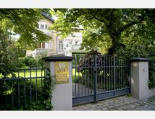 Ein zweigeschossiges Wohngebäude mit heller Fassade und großen Fenstern, umgeben von einem gepflegten Garten mit Rasen, Bäumen und blühenden Sträuchern. Im Vordergrund ein schmiedeeisernes Tor mit einem Briefkasten in einer Säule, auf dem die Hausnummer 25 zu sehen ist.